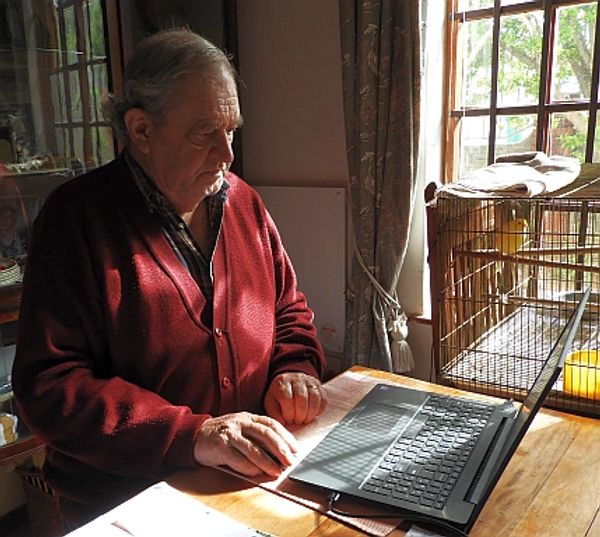 Elderly man using a laptop at a sunlit table near a birdcage.
