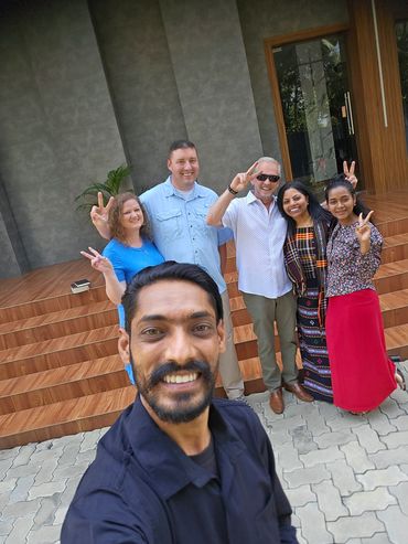 Group selfie with six people smiling and making peace signs outdoors.