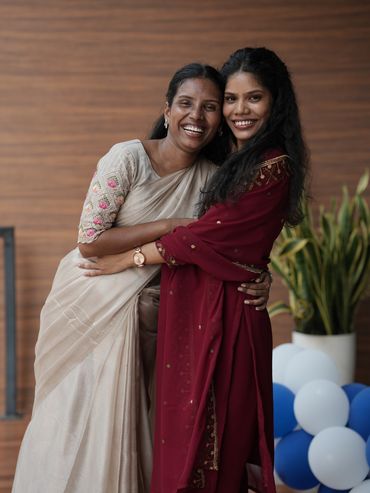 Two women in traditional attire happily embracing indoors.