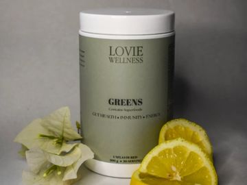Container of Lovie Wellness Greens supplement with sliced citrus and flowers.