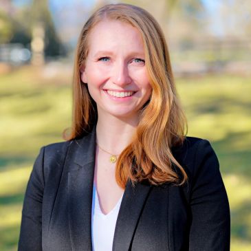 Woman with blonde hair smiling outdoors in a black blazer.