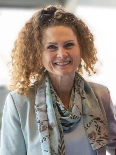 Smiling woman with curly hair wearing a light blue blazer and patterned scarf.