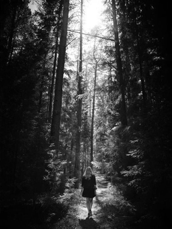 A lone woman walks through a sunlit forest path in black and white.