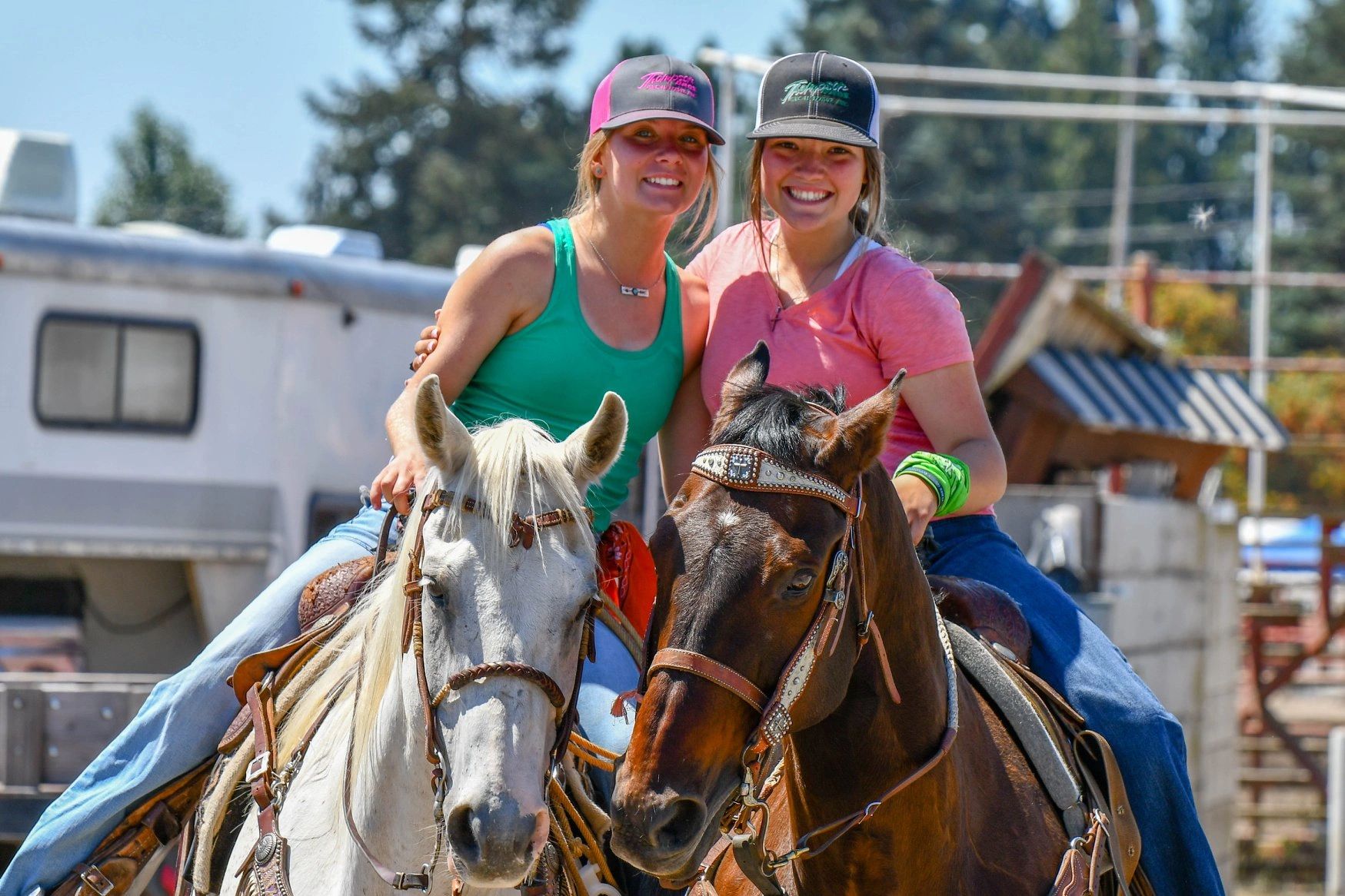 Two women smiling while riding horses outdoors on a sunny day.