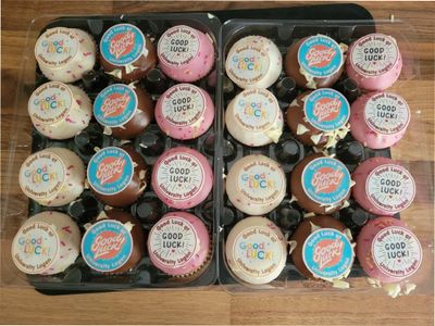 Assorted cupcakes with colorful 'Good Luck' and 'Good Luck Sweet' toppers in plastic trays.