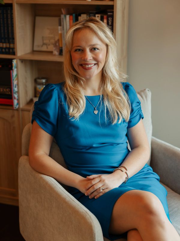 Smiling woman in blue dress sitting comfortably in an armchair indoors.