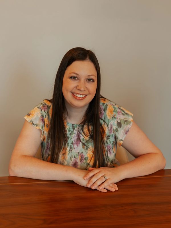 Smiling woman with long dark hair in a floral blouse sitting at a wooden table.