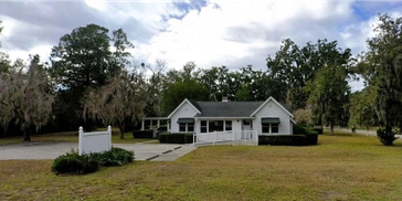 A small white house surrounded by trees and an open lawn under a cloudy sky.