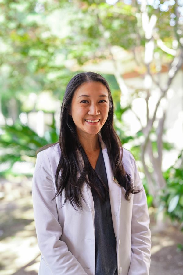 Smiling female doctor in white coat outdoors with greenery in background.
