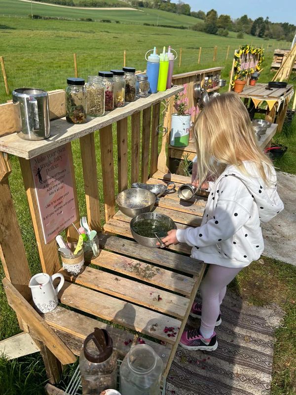 A child playing at an outdoor mud kitchen with jars of herbs and utensils.
