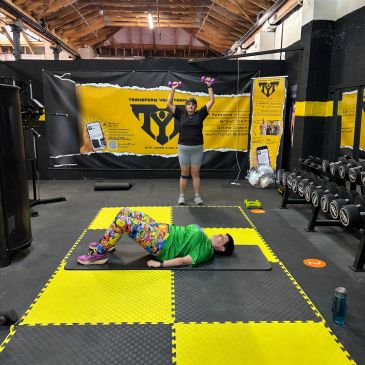 Two people exercising in a gym with yellow and black equipment.
