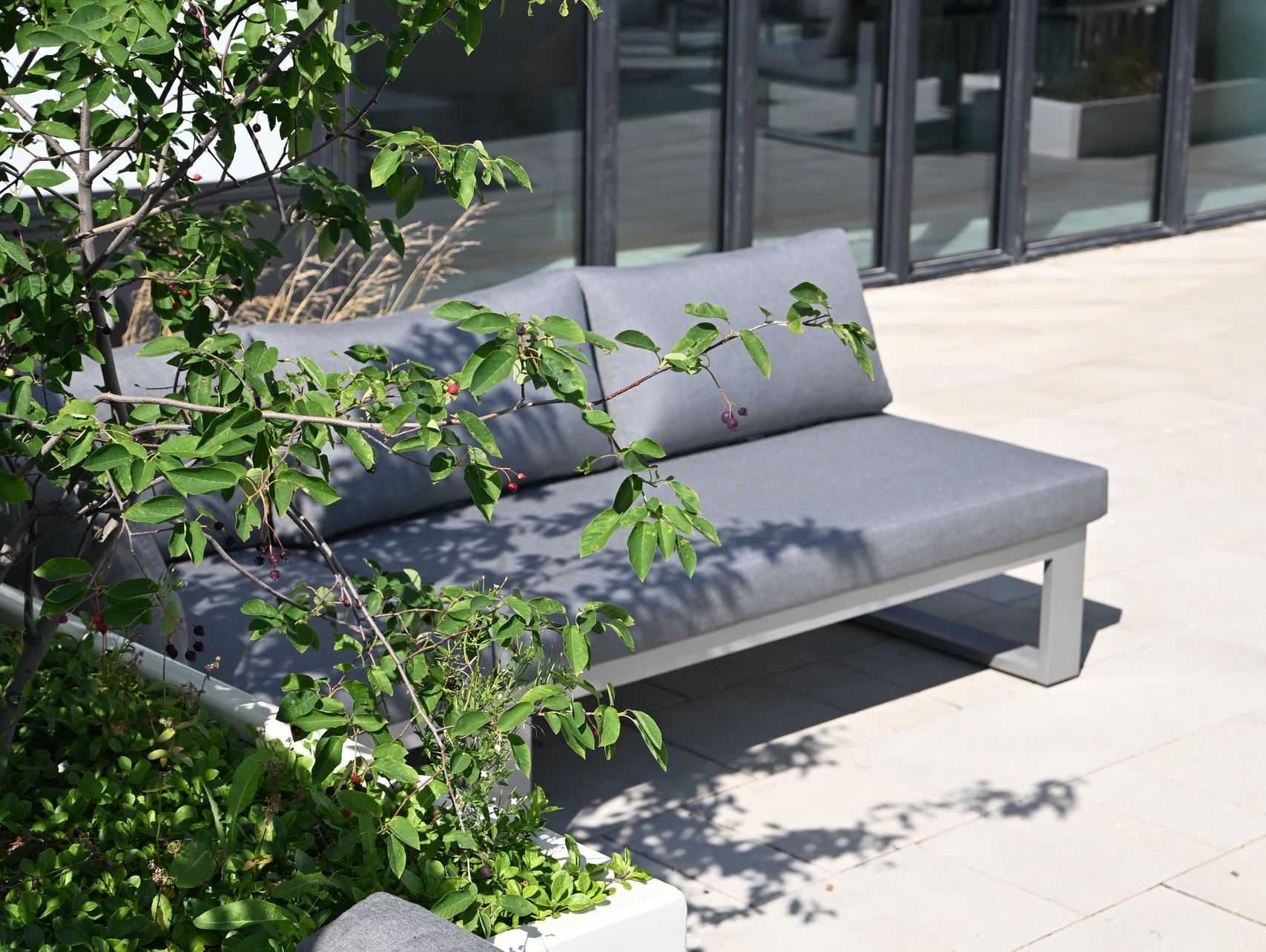 Gray outdoor sofa near green plants and glass windows.