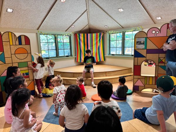 Children listening to a story read by a boy on a small stage in a colorful room.