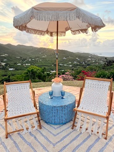 Two woven chairs and a table with an umbrella overlooking a scenic hillside at sunset.