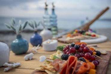 Close-up of a charcuterie board with fruits and crackers on a beach table.