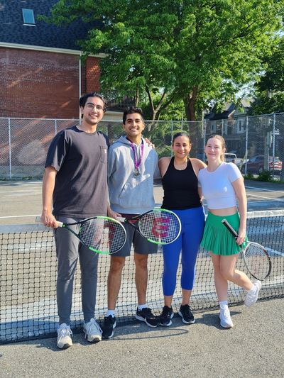 Four young adults posing with tennis rackets on a sunny tennis court.