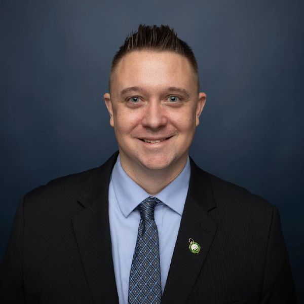 Smiling man in a suit and patterned tie against a dark blue background.