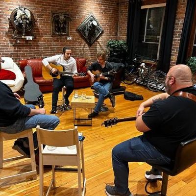 A group of songwriters sitting in a circle. Aiden and another songwriter are playing their guitars.