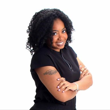 Confident woman with curly hair smiling, arms crossed, wearing black top with tattoo on arm.