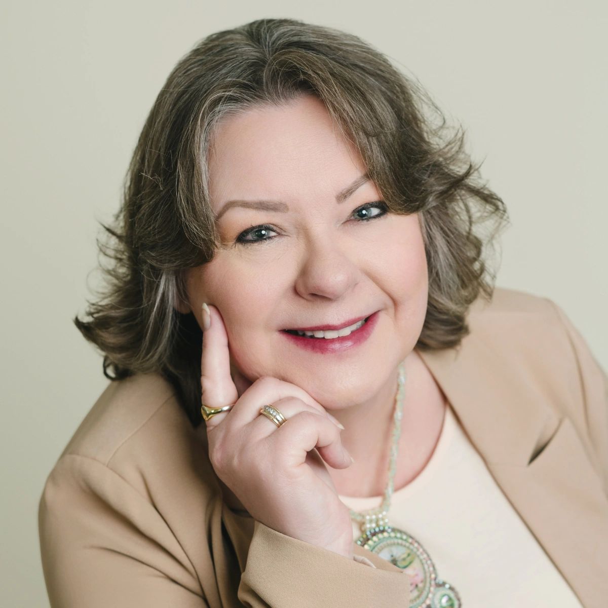 Smiling middle-aged woman with gray hair, wearing a beige blazer and a beaded necklace.