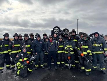 Group of firefighters posing in front of an overturned car under a cloudy sky.