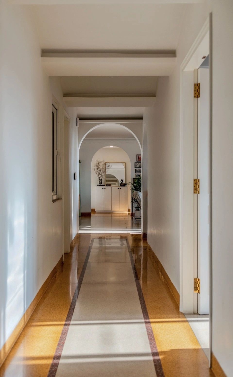 Sunlit hallway with archways and polished floor leading to a decorated cabinet.