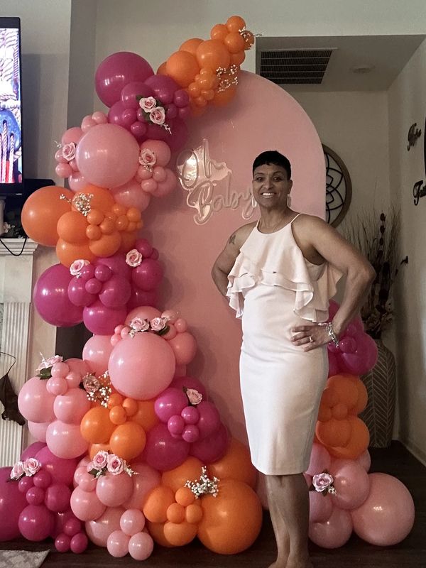 Woman in a white dress poses beside a vibrant balloon arch for a baby shower.