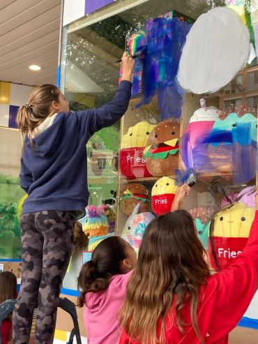 Kids painting a shop window with colorful food designs.