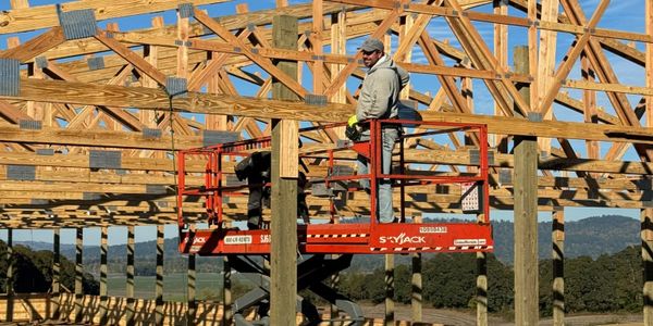 A worker on a scissor lift assembling wooden trusses outdoors.