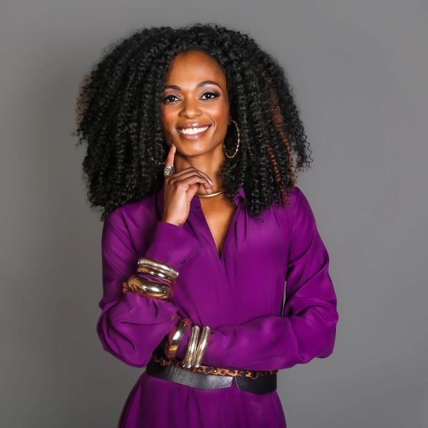 Confident woman with voluminous curly hair in a purple dress smiling.