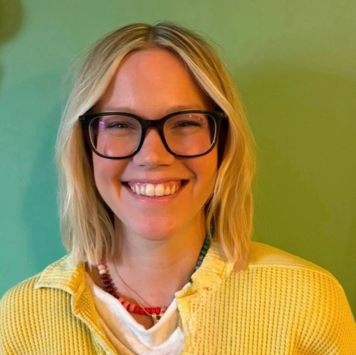 Smiling woman with glasses wearing a yellow sweater against a green background.