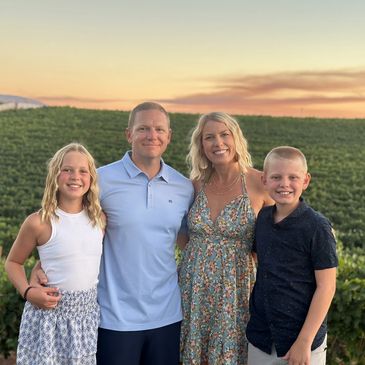 Smiling family of four posing in front of a vineyard at sunset.