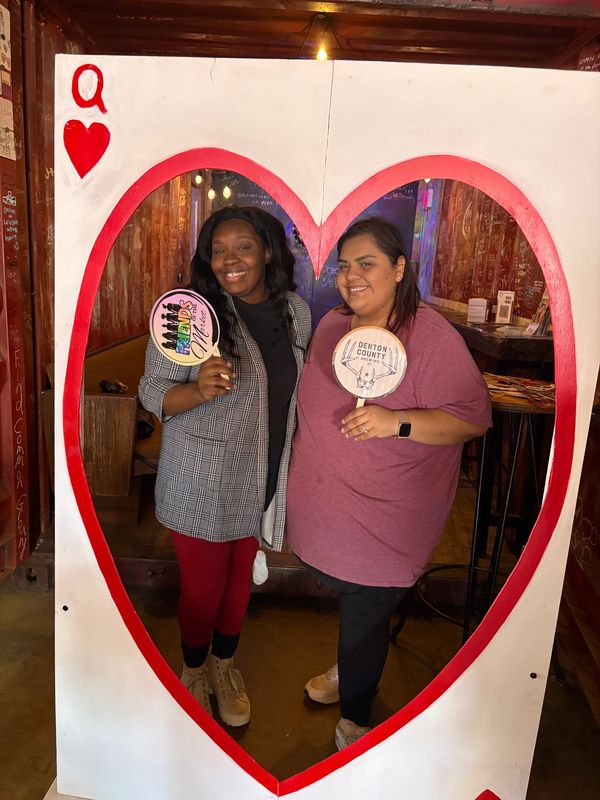 Two women posing inside a Queen of Hearts card cutout at a cozy venue.