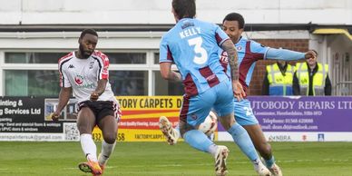 A soccer player in white kicks the ball while two opponents in blue defend.