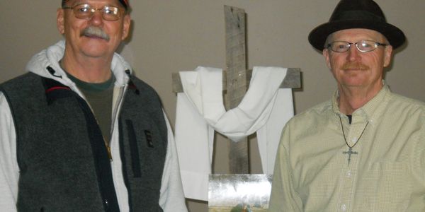 Two men stand beside a wooden cross draped with a white cloth