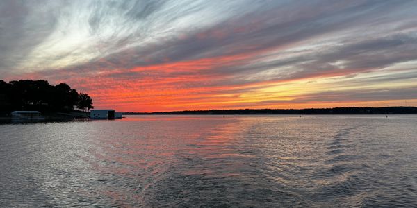 Vibrant sunset over a calm lake with silhouetted trees and buildings.