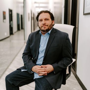 Man with beard sitting in an office hallway on a chair wearing a dark grey suit and blue shirt