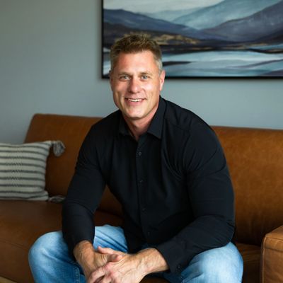 Smiling man in black shirt sitting on a brown leather couch.