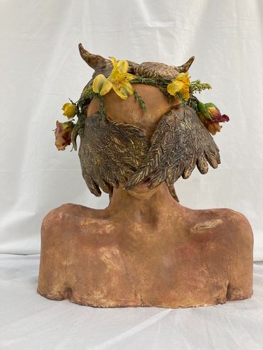 Sculpture of a person with bird wings covering the eyes and flowers on the head.