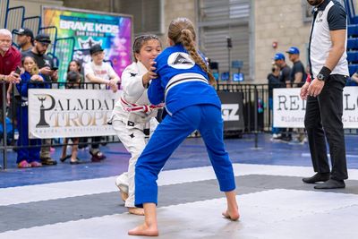 Two young girls competing in a Brazilian Jiu-Jitsu match with a referee nearby.