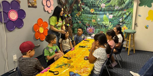 A woman teaches a group of children seated around a colorful table with a bee-themed tablecloth.