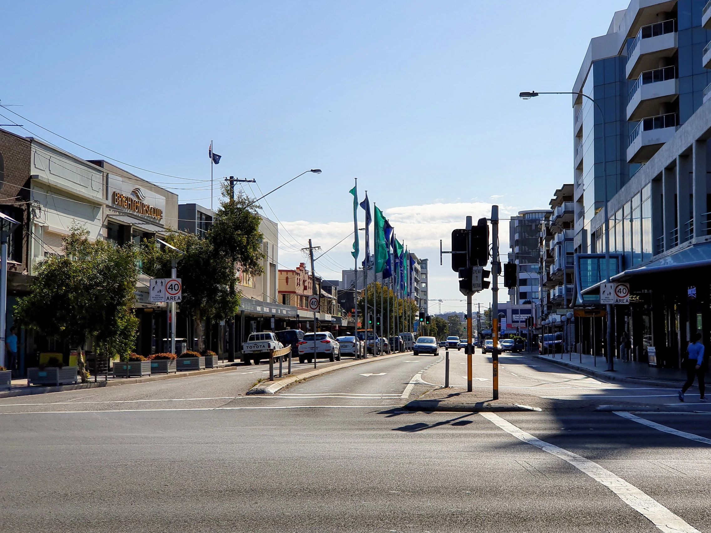 A sunny city street with cars and pedestrians near buildings and flags.