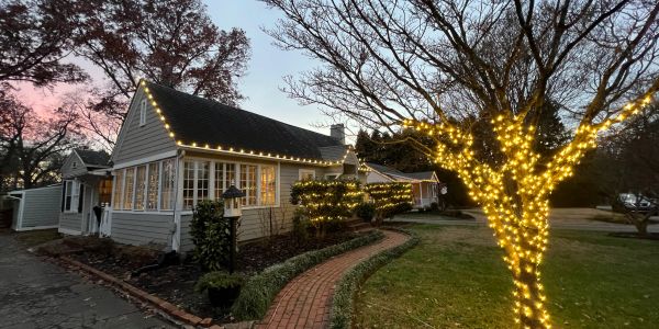 Cozy house decorated with warm string lights on trees and roof edges at dusk.