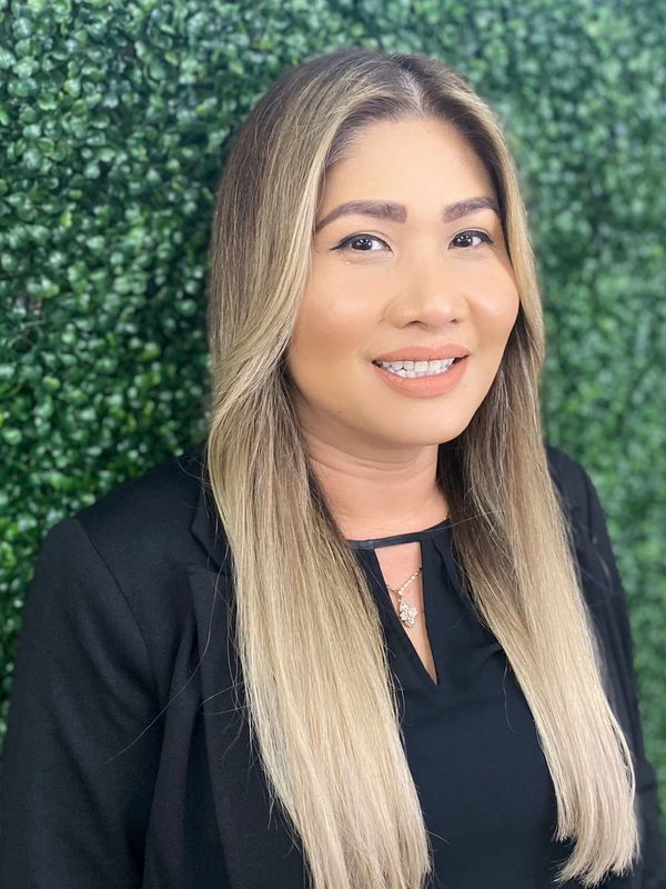 Professional woman with long blonde hair and black blazer smiling against a green leafy background.