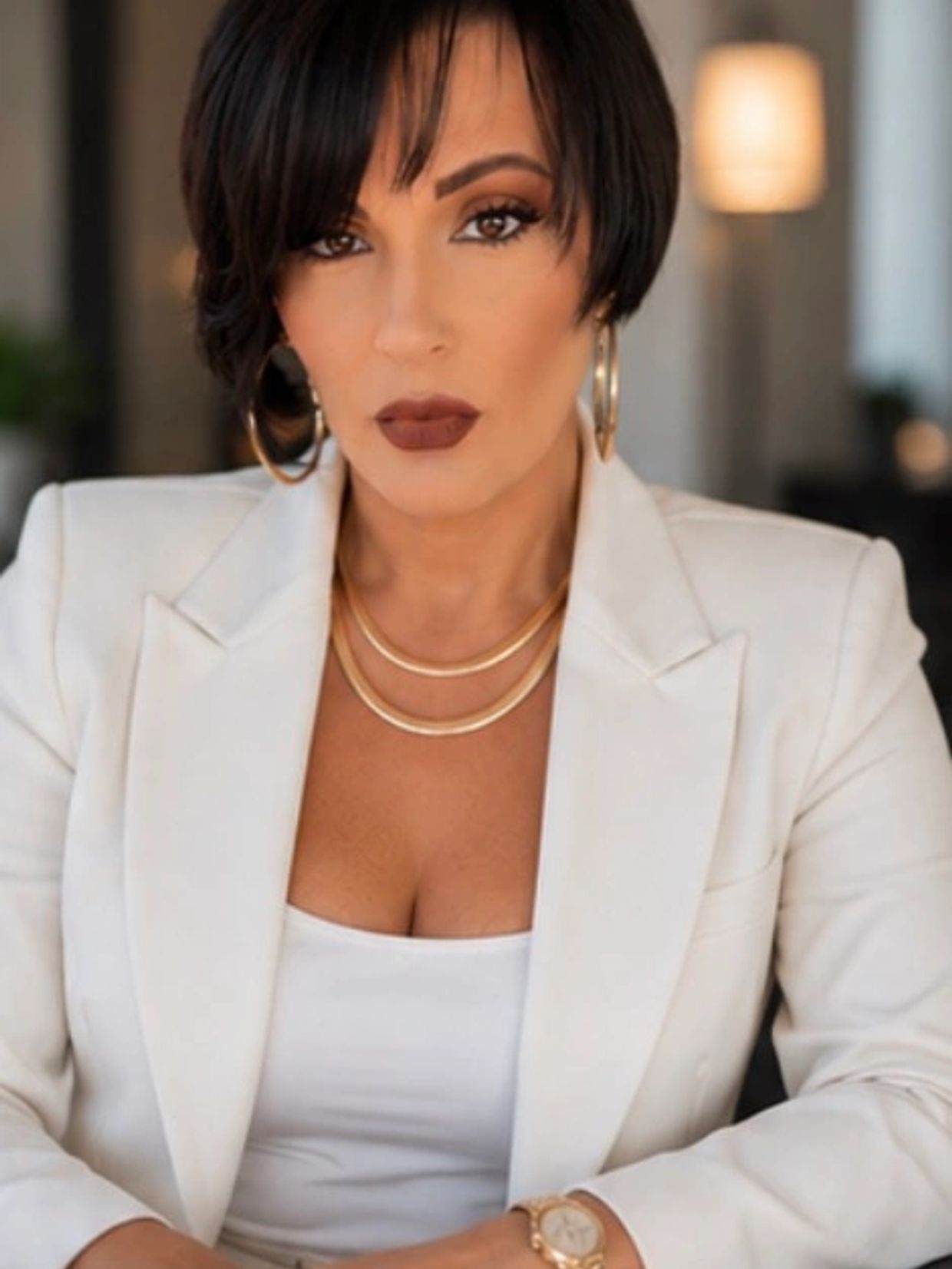 Confident woman in a white blazer with gold jewelry and bold makeup.