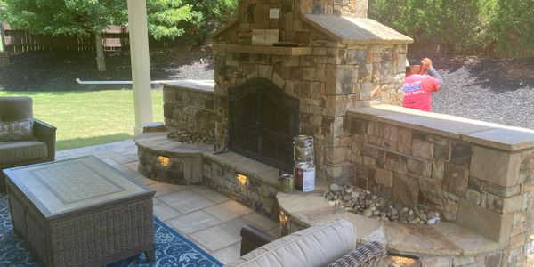 Cozy outdoor stone fireplace with seating and a wicker table on a patio.