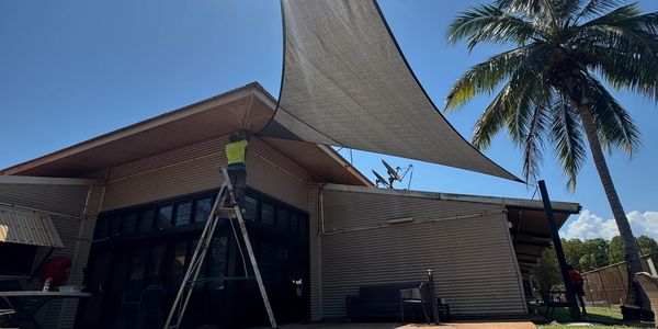 Man on ladder installing a shade sail on a sunny day beside a palm tree.