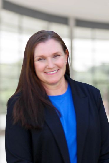 Smiling woman with long brown hair wearing a blue shirt and black jacket.