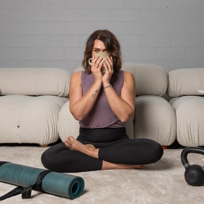 Woman in workout clothes sitting cross-legged, drinking from a mug indoors.