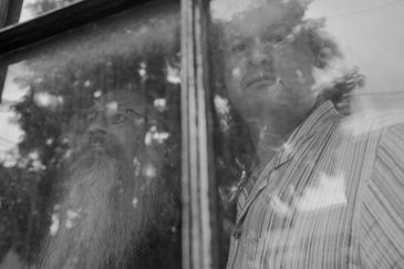 Black & white photo of Aris Karabelas & Mike Magee of band Nom De Plume looking out rustic window.